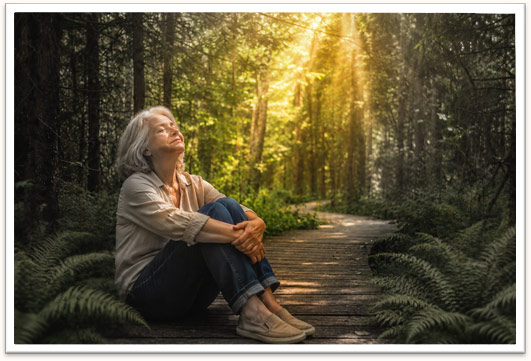 An older woman sits peacefully on a wooden path in a sunlit forest, symbolizing rest and healing